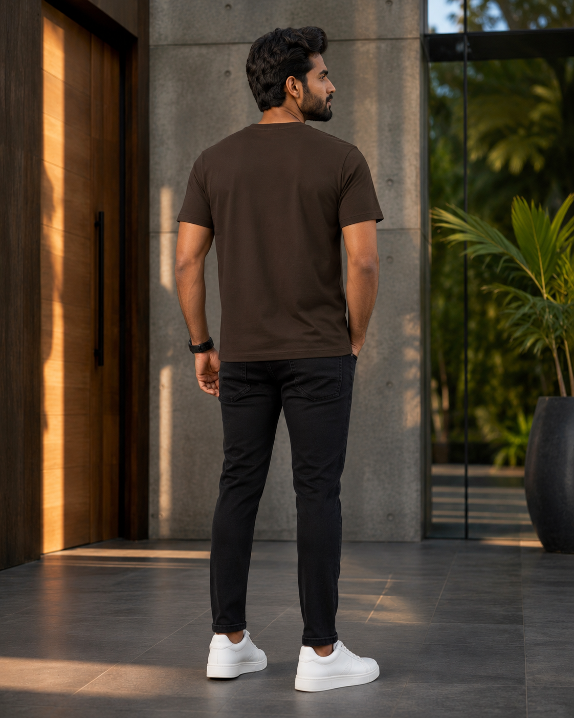 Back view of a model wearing a premium brown cotton t shirt with black slim fit pants and white sneakers outdoors.