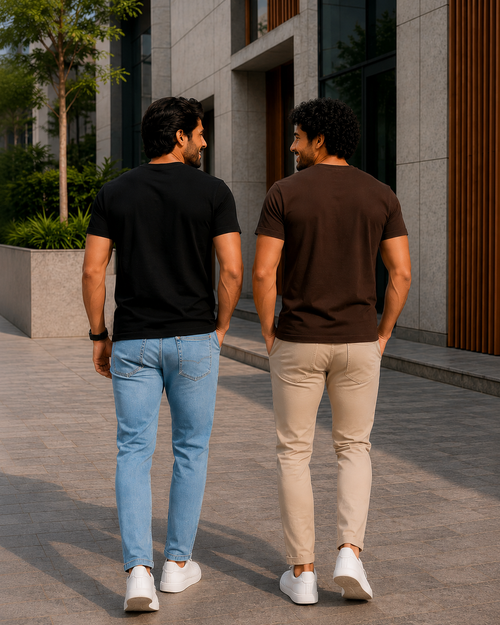 Back view of two men wearing plain crew neck T-shirts for men one in black with light blue jeans and the other in dark brown with beige chinos walking outdoors in a modern urban setting with white sneakers.