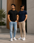 Two men standing in a modern urban setting wearing plain crew neck T-shirts for men one in black with light blue jeans and the other in navy blue with beige chinos paired with white sneakers for a clean casual look.