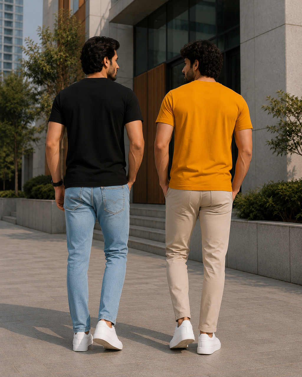 Back view of two men wearing plain crew neck T-shirts one in black with light blue jeans and the other in orange t shirt for men with beige chinos walking outdoors in a modern urban setting with white sneakers.