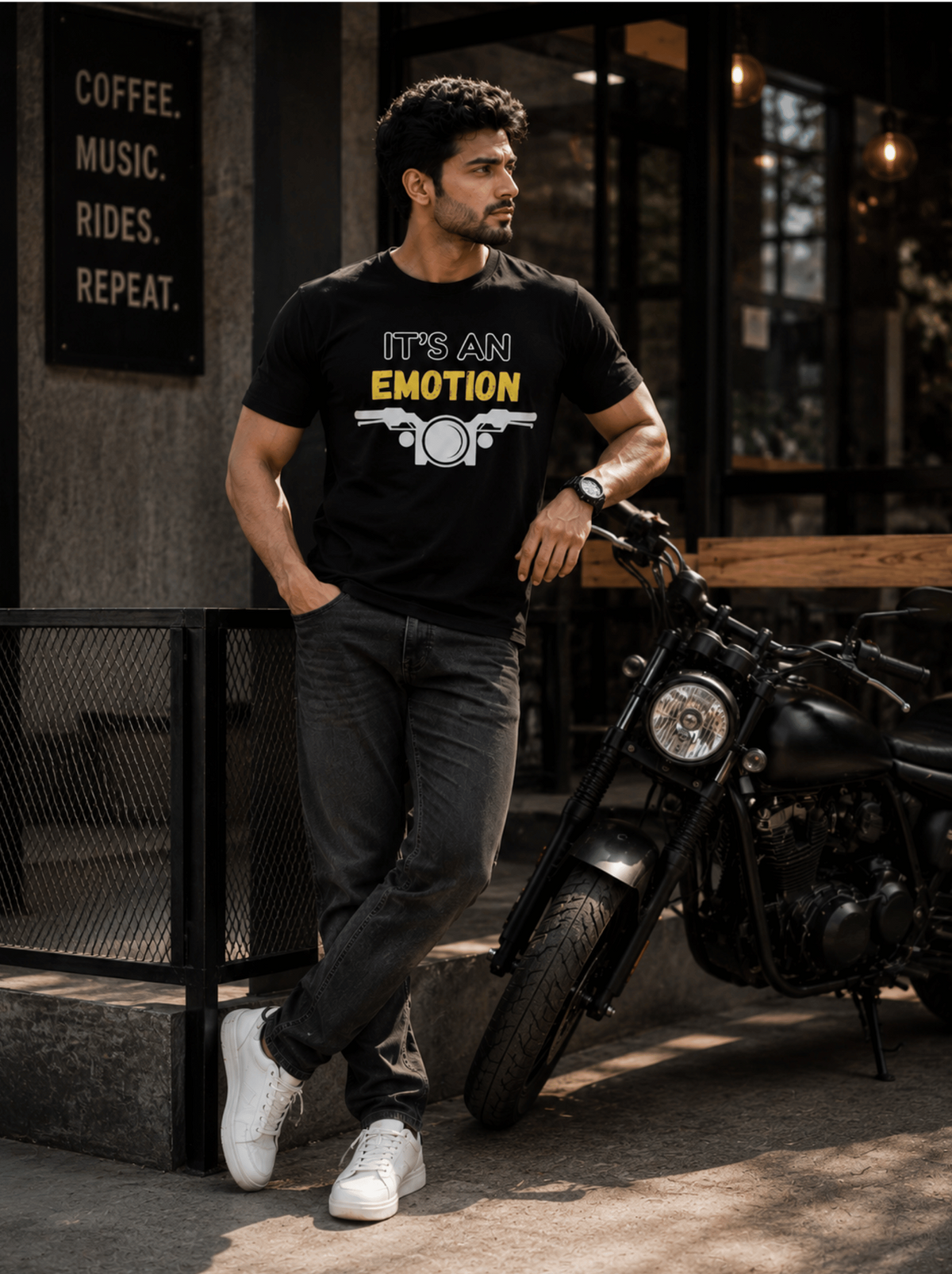 A man wearing a black graphic t-shirt with the text 'IT'S AN EMOTION', standing next to a black motorcycle in front of a cafe.