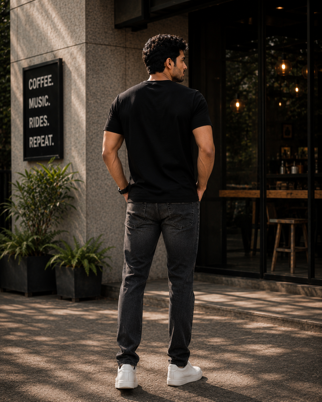 A man standing outdoors in front of a coffee shop, viewed from behind. He is wearing a black short sleeve t-shirt, dark gray jeans, and white sneakers.