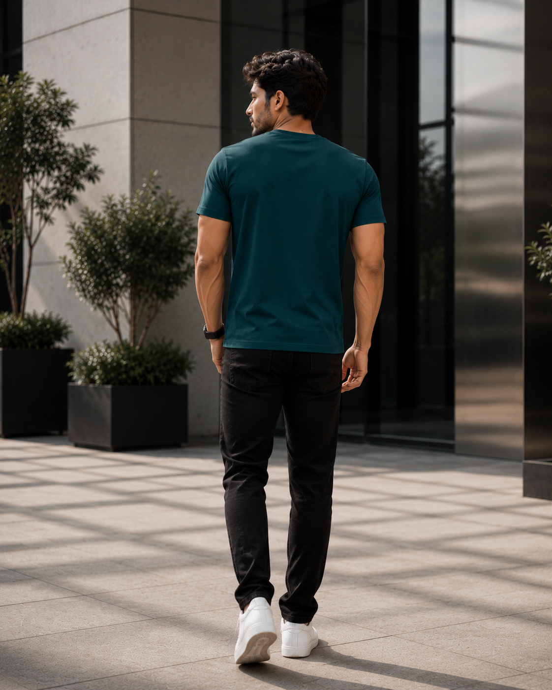 A man viewed from behind wearing a dark teal short sleeve t-shirt, black pants, and white sneakers, walking outdoors in front of a modern building with glass windows and potted plants.