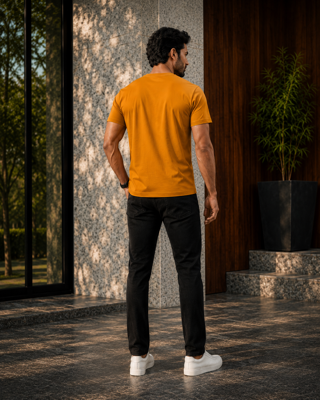 Back view of a model wearing a premium orange t shirt for men with black pants and white sneakers outside a modern building.