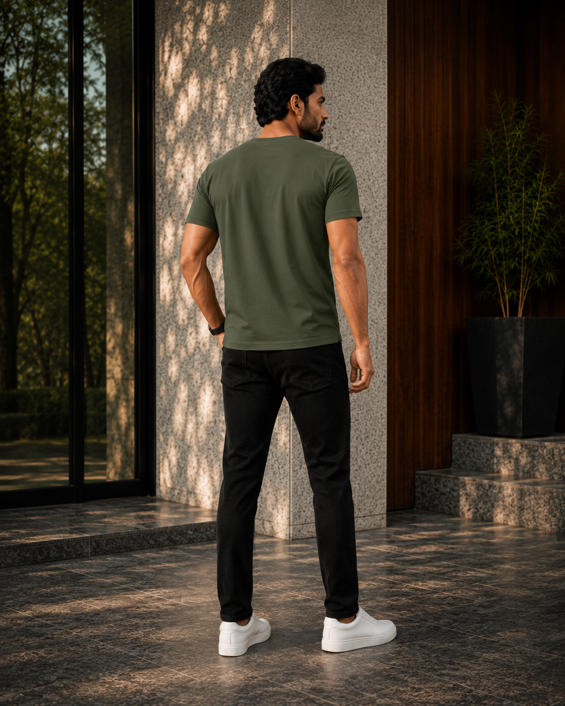 A man viewed from behind wearing an olive green short sleeve t shirt for men, black slim-fit pants, and white sneakers, standing on a stone patio in front of a modern building.