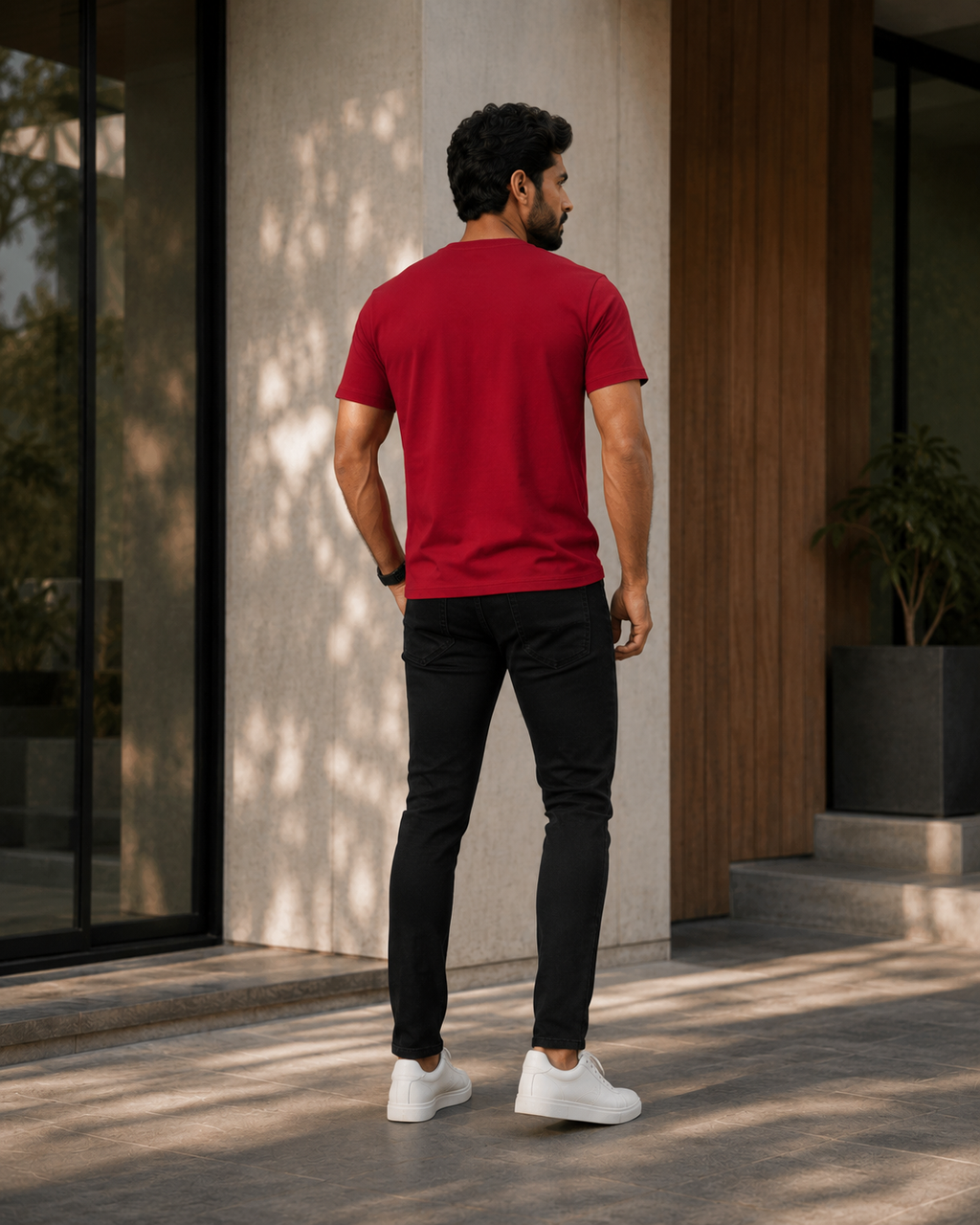 Back view of a model wearing a premium plain red t shirt for men with black slim fit jeans and white sneakers outside a modern building.